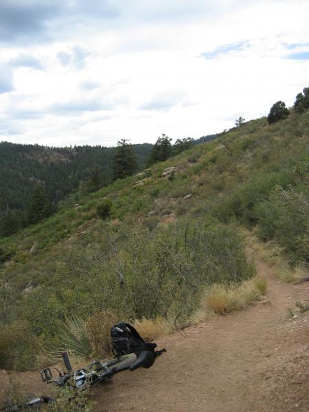 A scenic view of a mountainous trail surrounded by greenery, with a bicycle lying on the ground and a backpack nearby. The path winds through a hillside dotted with shrubs and trees under a cloudy sky. Waldo Canyon mountain bike trail.