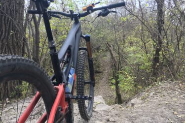 A mountain bike positioned on a rocky trail, surrounded by trees and greenery, with a winding path visible in the background. The bike is angled to show its wheels and handlebars, highlighting its features against the natural setting. Palos Forest Preserve mountain bike trail.