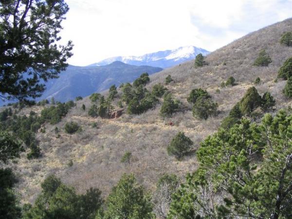 A panoramic view of a mountainous landscape featuring a mix of trees and sparse vegetation, with a snow-capped mountain peak visible in the background. The terrain is hilly, indicating a natural setting under a partly cloudy sky. Waldo Canyon mountain bike trail.