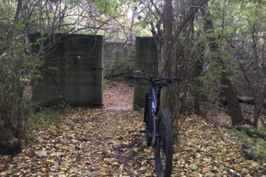 A mountain bike is parked on a leaf-covered path leading to a partially obscured concrete structure surrounded by trees and dense foliage. The scene is tranquil and slightly overgrown, suggesting an adventurous or exploratory atmosphere. Palos Forest Preserve mountain bike trail.