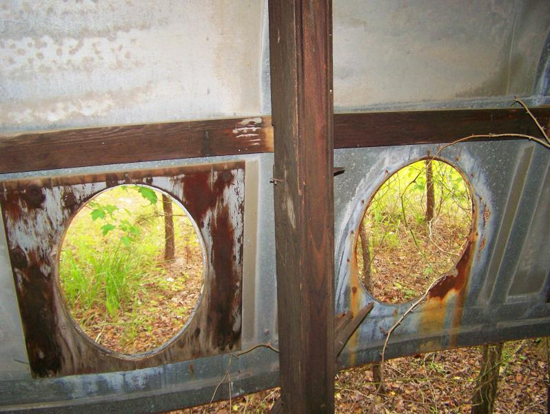 An interior view of an aged and weathered metal structure featuring two circular windows framed by wooden supports, surrounded by greenery visible through the openings. The walls show signs of rust and wear. Iron Legs Trail mountain bike trail.