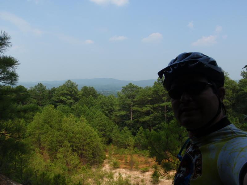 A person wearing a bicycle helmet stands in front of a scenic view of rolling hills and dense green forests under a clear blue sky. Iron Legs Trail mountain bike trail.