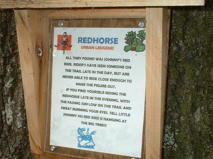 A sign attached to a tree, titled "Redhorse Urban Legend." The sign features illustrations and text describing a local legend about sightings of a boy named Johnny and his red bike. The text warns riders about encounters with the mysterious bike in the evening. Killer Three Loop mountain bike trail.
