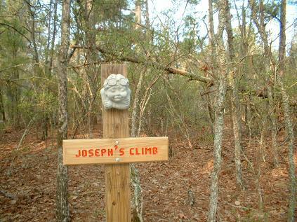 Wooden trail sign reading "Joseph's Climb," featuring a decorative stone face at the top, surrounded by a wooded area with trees and leaves on the ground. Killer Three Loop mountain bike trail.
