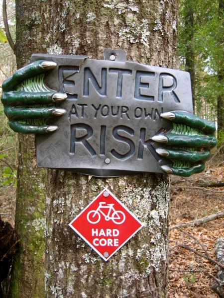 A tree trunk features a sign that reads "ENTER AT YOUR OWN RISK," supported by green, claw-like hands gripping the edges. Below, there is a red diamond-shaped sign with a bicycle symbol and the words "HARD CORE," indicating a challenging trail or area. Killer Three Loop mountain bike trail.