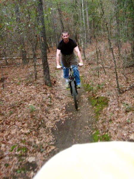 A person riding a mountain bike on a dirt trail in a wooded area, surrounded by trees and leaves. The cyclist is wearing a dark T-shirt and jeans, and is positioned in the foreground, approaching the camera. The scene is natural, showcasing the outdoors and recreational biking. Killer Three Loop mountain bike trail.