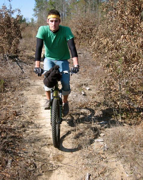 A cyclist wearing a green shirt, long black sleeves, and a yellow headband rides a mountain bike on a dirt trail surrounded by dry vegetation and trees. The cyclist has a focused expression, and a black garment is draped over the handlebars. Killer Three Loop mountain bike trail.