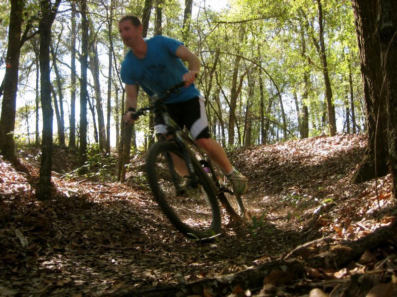 A mountain biker in a blue shirt navigates a twisting trail through a lush forest, surrounded by trees and fallen leaves, catching sunlight filtering through the canopy. Killer Three Loop mountain bike trail.