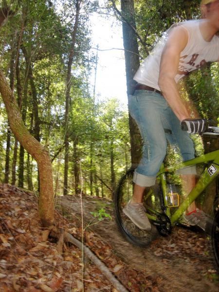 A person riding a green mountain bike on a trail in a wooded area, with trees and foliage in the background. The image captures the cyclist in motion, wearing casual outdoor clothing and gloves. Killer Three Loop mountain bike trail.