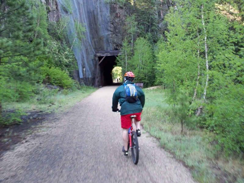 A person riding a bicycle down a gravel path surrounded by lush greenery, approaching a dark tunnel in the distance. The George S. Mickelson Trail mountain bike trail.