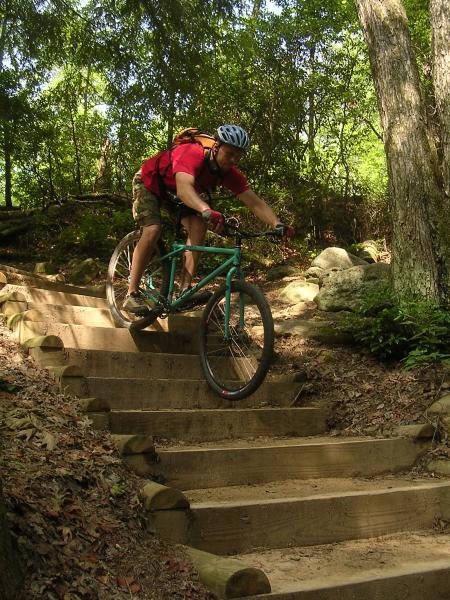 A person riding a mountain bike down a set of wooden stairs in a forested area. The rider is leaning forward, wearing a helmet and a red shirt, demonstrating balance and skill on the descent. Surrounding the stairs are trees and rocks, creating a natural outdoor environment. Chilhowee trail system mountain bike trail.