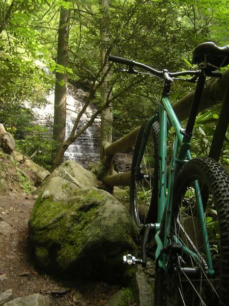 A mountain bike leaning against a large rock on a forest path, with a cascading waterfall visible in the background surrounded by lush greenery. Chilhowee trail system mountain bike trail.