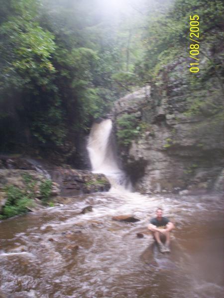 A man sitting on a rock in a river, with a waterfall cascading down rocky cliffs in the background, surrounded by lush green vegetation. The scene is slightly misty, suggesting a humid environment. The date "01/08/2005" is visible in the corner of the image. Chilhowee trail system mountain bike trail.