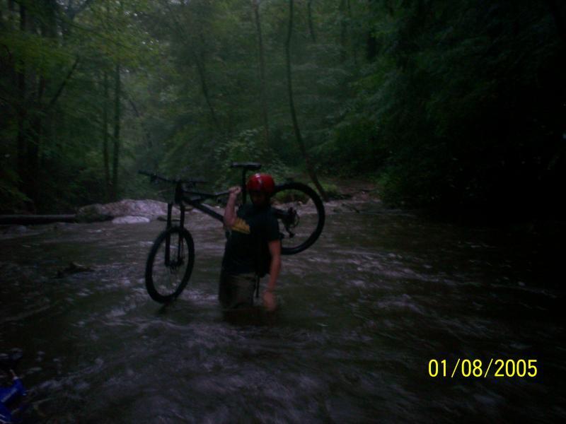 A person wearing a helmet stands in a flowing stream, carrying a mountain bike above the water. Lush green trees and a misty atmosphere surround the scene, indicating a natural, outdoor setting. Chilhowee trail system mountain bike trail.