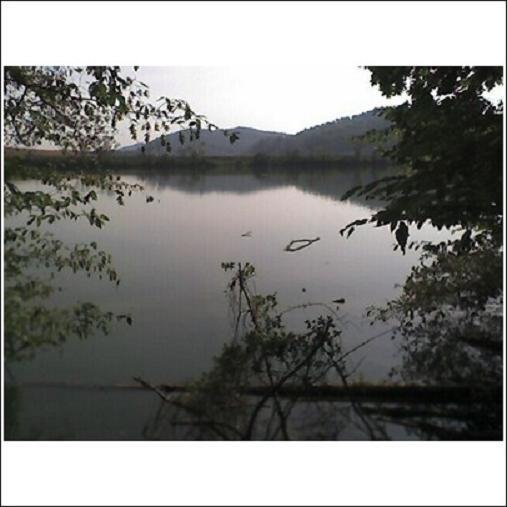 A tranquil lakeside scene surrounded by trees and mountains in the background. The calm water reflects the overcast sky, creating a peaceful atmosphere. Haw Ridge Park mountain bike trail.