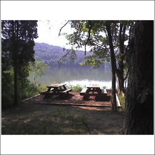 A serene lakeside scene featuring two picnic tables set on a sandy patch surrounded by greenery. In the background, calm water reflects the gentle slopes of distant hills under a clear sky. The area is shaded by trees, creating a peaceful outdoor setting. Haw Ridge Park mountain bike trail.