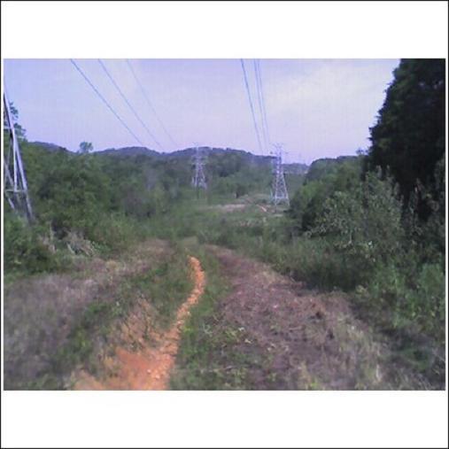 A dirt path leading through a grassy area with power lines running overhead, surrounded by trees and rolling hills in the background. The sky is partially cloudy. Haw Ridge Park mountain bike trail.