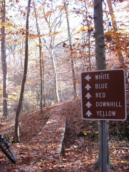 Trail sign in a wooded area directing hikers and bikers to different paths marked by colors: white, blue, red, downhill, and yellow. The ground is covered with fallen leaves, and a wooden bridge leads along the trail. The scene captures the beauty of autumn in a forested setting. Montgomery Bell State Park Mtb Trail mountain bike trail.