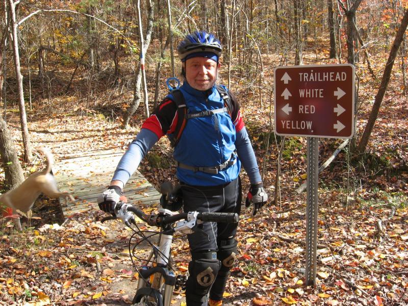 A middle-aged man in biking gear, including a blue shirt and a helmet, stands next to his mountain bike at a trailhead in a forested area. A sign behind him indicates different trail directions: white, red, and yellow. The ground is covered with fallen leaves, and a blurred dog can be seen in the background. Montgomery Bell State Park Mtb Trail mountain bike trail.