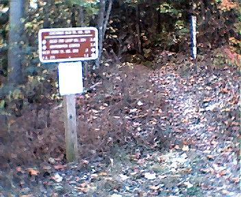 A trail sign marking distances to various destinations, located at a wooded path. The area has fallen leaves and shrubbery surrounding the sign. Tanasi Trail System mountain bike trail.