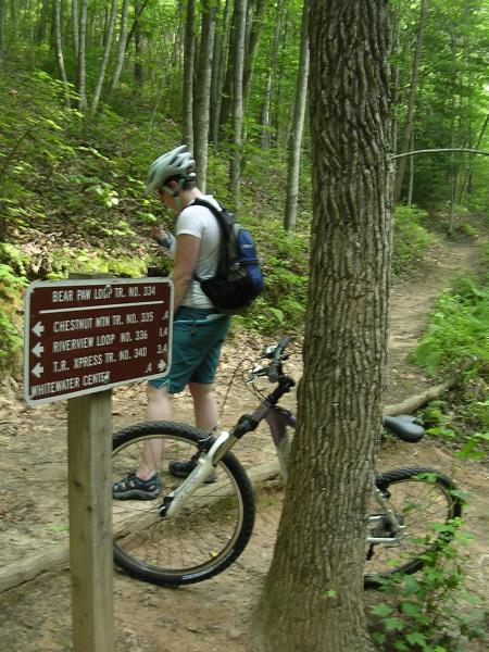 A person wearing a helmet and a backpack is standing next to a wooden trail sign in a wooded area. The sign lists various trail names and numbers. A mountain bike is leaned against the sign, and a dirt path leads into the greenery of the forest. Sunlight filters through the trees, creating a vibrant outdoor scene. Tanasi Trail System mountain bike trail.