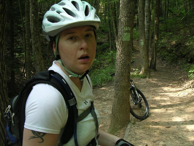 A person wearing a white helmet and a backpack stands on a mountain biking trail surrounded by trees. They appear to be catching their breath, with a mountain bike partially visible behind them on the path. The environment is lush and green, indicating a wooded area. Tanasi Trail System mountain bike trail.