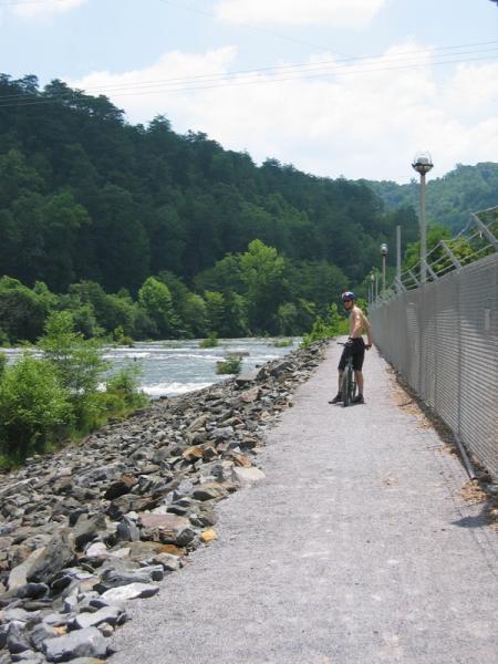 A shirtless cyclist stands on a gravel path alongside a river, surrounded by lush green trees and hills. The cyclist is holding a bike and looking towards the water, while a chain-link fence lines the path. The scene is bright and captures a sunny day outdoors. Tanasi Trail System mountain bike trail.