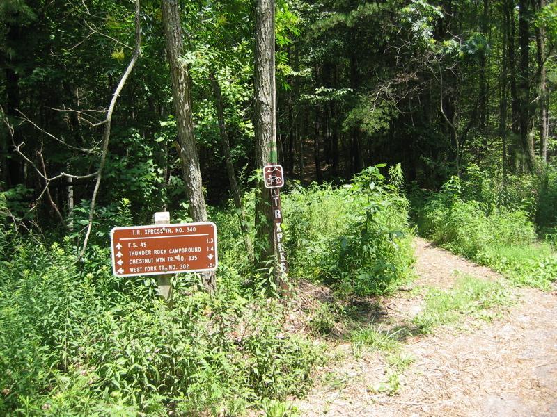 Trail intersection sign indicating various hiking routes, surrounded by dense greenery and trees. The sign lists distances to Thunder Rock Campground, Chestnut Mountain, and West Fork Trail. A narrow path leads into the forest. Tanasi Trail System mountain bike trail.