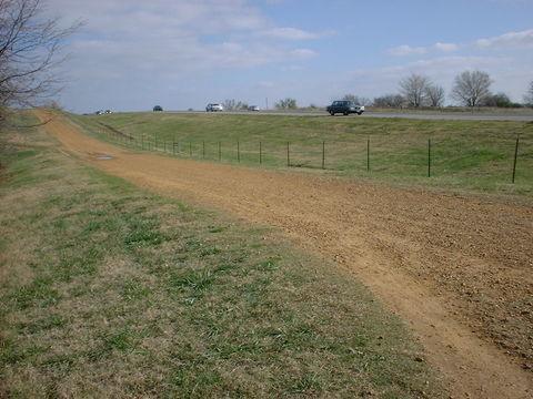 A dirt road leading up a small hill, with a gravel surface and grassy areas on either side. In the background, a paved highway can be seen with several vehicles passing by. The sky is partly cloudy, and there are a few trees in the distance. Tour de Wolf mountain bike trail.