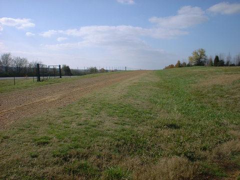 A grassy hillside with a dirt path, surrounded by a wire fence, under a partly cloudy blue sky. The landscape is open with a mix of grass and bare patches on the slope. Tour de Wolf mountain bike trail.