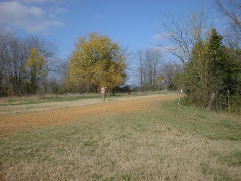 A gravel pathway leading to a signpost, surrounded by grassy fields and sparse trees, with a clear blue sky overhead and a few clouds. Tour de Wolf mountain bike trail.