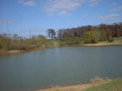 A peaceful landscape featuring a calm, serene lake surrounded by trees and greenery under a partially cloudy sky. The water reflects the colors of the surrounding nature, creating a tranquil setting. Tour de Wolf mountain bike trail.