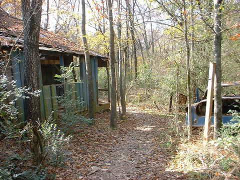 A narrow dirt path winding through a forested area, leading to a weathered, abandoned building on the left. The building, covered in peeling blue paint, has a partially open door and is surrounded by trees and underbrush. On the right side of the path, an old, rusted vehicle is partially visible, framed by fallen leaves and greenery. The scene is set in autumn, with warm light filtering through the trees. Tour de Wolf mountain bike trail.