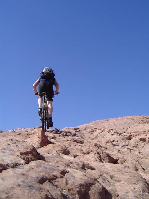 A mountain biker ascends a rocky terrain under a clear blue sky, showcasing the challenge of off-road cycling. The cyclist, dressed in black shorts and a backpack, is focused on navigating the uneven surface as they pedal upward. Slickrock mountain bike trail.