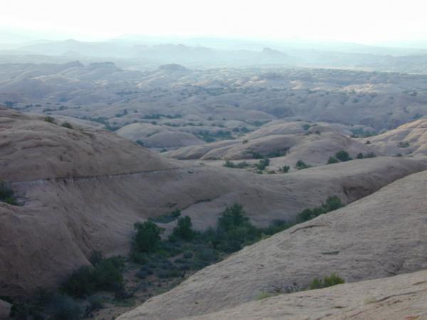 A panoramic view of a rocky landscape featuring rolling, rounded hills and sparse vegetation, with distant mountains under a soft, hazy sky. The terrain is predominantly beige and light brown, indicating rugged, arid conditions. Slickrock mountain bike trail.