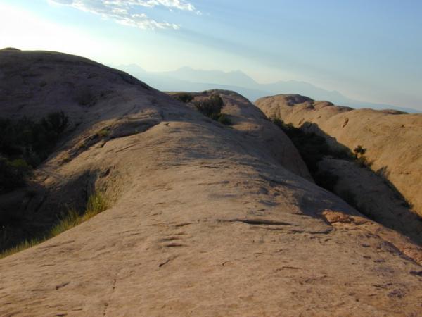 A sweeping view of a rocky landscape with smooth, undulating terrain and patches of greenery. In the background, distant mountains are silhouetted against a sky with soft clouds and warm sunlight casting gentle shadows on the rocks. Slickrock mountain bike trail.