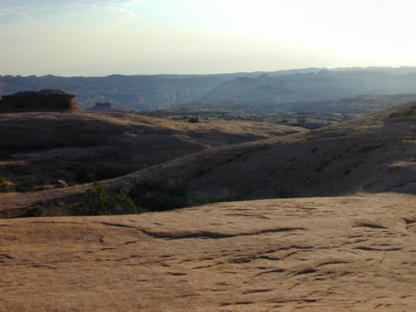 A panoramic view of a rocky desert landscape under a soft evening light, showcasing rolling hills and distant mountains. The foreground features smooth, flat rock surfaces, while sparse vegetation dots the terrain. The sky is partly cloudy, hinting at sunset. Slickrock mountain bike trail.