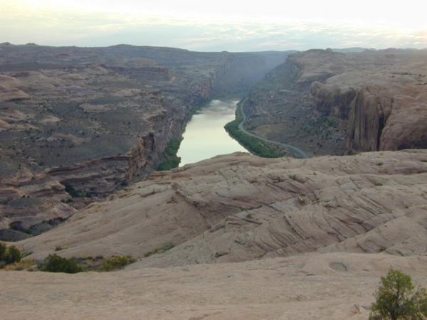 A panoramic view of a rugged canyon landscape featuring a winding river surrounded by steep rock formations and layered geological structures. The scene is bathed in soft, ambient light, capturing the serene beauty of nature. Slickrock mountain bike trail.