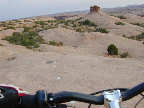 A view from the perspective of a mountain biker, showing rocky terrain with scattered vegetation and a prominent rock formation in the distance. The handlebars of the bike are visible in the foreground, and the landscape features smooth, rounded surfaces and a blue sky. Slickrock mountain bike trail.