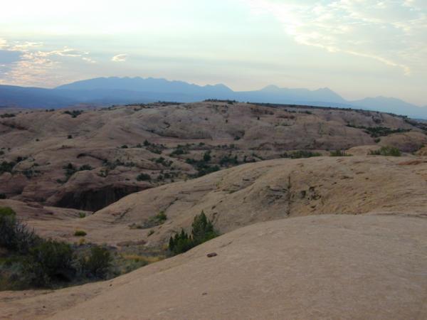 Landscape view of rolling sandstone formations under a cloudy sky, with distant mountains visible in the background. The terrain features sparse vegetation and rugged terrain, showcasing the natural beauty of the area. Slickrock mountain bike trail.