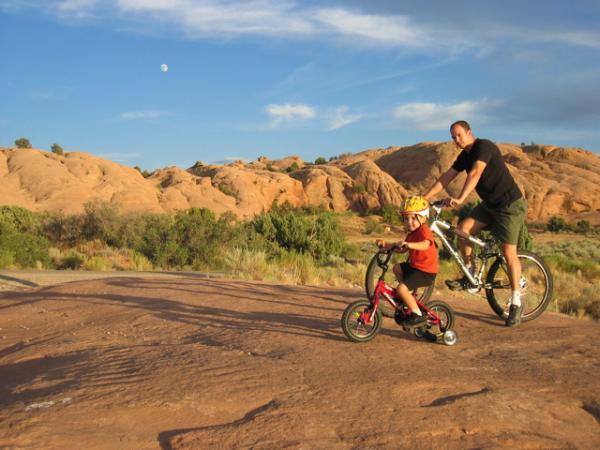 A man riding a bike alongside a child on a small scooter, both enjoying an outdoor adventure on a rocky path. The landscape features red rock formations and greenery under a blue sky with a visible moon. Slickrock mountain bike trail.