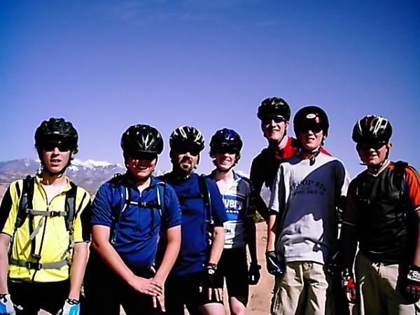 A group of eight individuals, diverse in age and gender, stand together outdoors wearing biking helmets and athletic clothing. They are smiling and posing for the camera against a backdrop of mountains and a clear blue sky. Some are wearing gloves, and the environment suggests a cycling or outdoor adventure. Slickrock mountain bike trail.