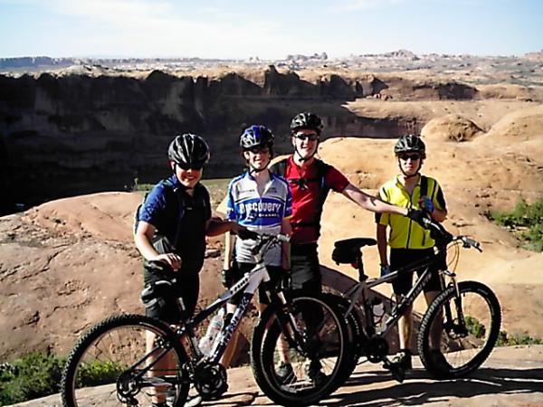 A group of four mountain bikers standing on a rocky overlook, posing next to their bikes with a vast canyon landscape in the background. They are wearing helmets and cycling attire, smiling and enjoying a sunny day outdoors. Slickrock mountain bike trail.
