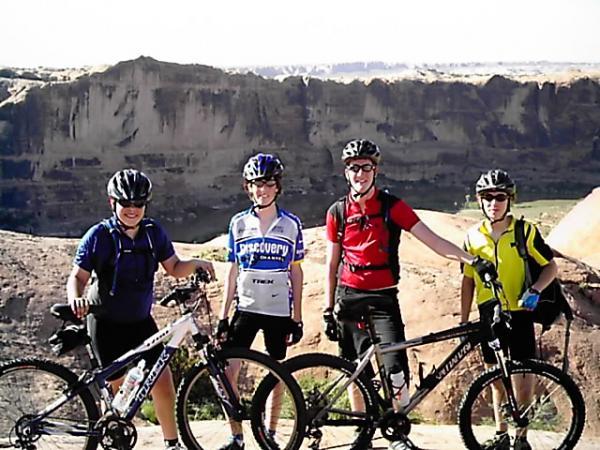 Four mountain bikers posing together with their bikes against a scenic backdrop of rocky cliffs and a clear sky. They are wearing helmets, cycling jerseys, and shorts, showcasing a range of colors. The landscape behind them features a dramatic canyon view. Slickrock mountain bike trail.