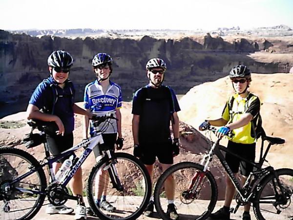 Four cyclists are standing with their mountain bikes on a rocky outcrop, overlooking a vast canyon landscape. The group is wearing helmets and biking gear, with two individuals in blue jerseys, one in black, and another in yellow. The sun is shining, highlighting the rugged terrain and distant cliffs in the background. Slickrock mountain bike trail.