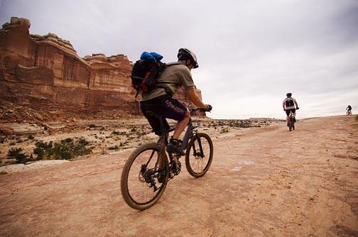 Mountain bikers riding on a rocky trail with dramatic cliffs in the background under a cloudy sky. Slickrock mountain bike trail.