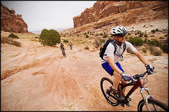 A group of mountain bikers navigating a rocky trail in a desert landscape. The foreground features a cyclist wearing a helmet and a hydration pack, focused on the terrain ahead. In the background, two other bikers can be seen riding along the rugged path, with red rock formations and sparse vegetation surrounding them. Slickrock mountain bike trail.