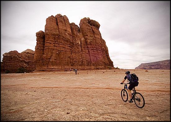 A mountain biker rides across a barren landscape with towering rock formations in the background, under a cloudy sky. Another cyclist can be seen in the distance. The scene captures the beauty of a desert environment. Slickrock mountain bike trail.
