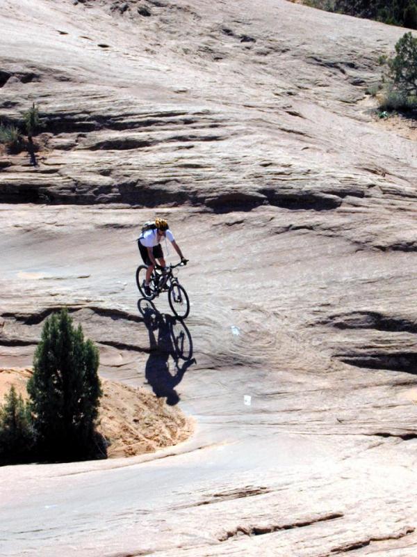 A mountain biker riding on smooth, rocky terrain with a slight incline, surrounded by sparse vegetation under a clear blue sky. The biker casts a shadow on the rock surface as they navigate the path. Slickrock mountain bike trail.