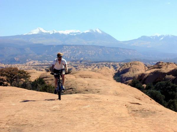 A person riding a mountain bike on a rocky terrain, with a backdrop of snow-capped mountains under a clear blue sky. The landscape features smooth rock formations and sparse vegetation. Slickrock mountain bike trail.